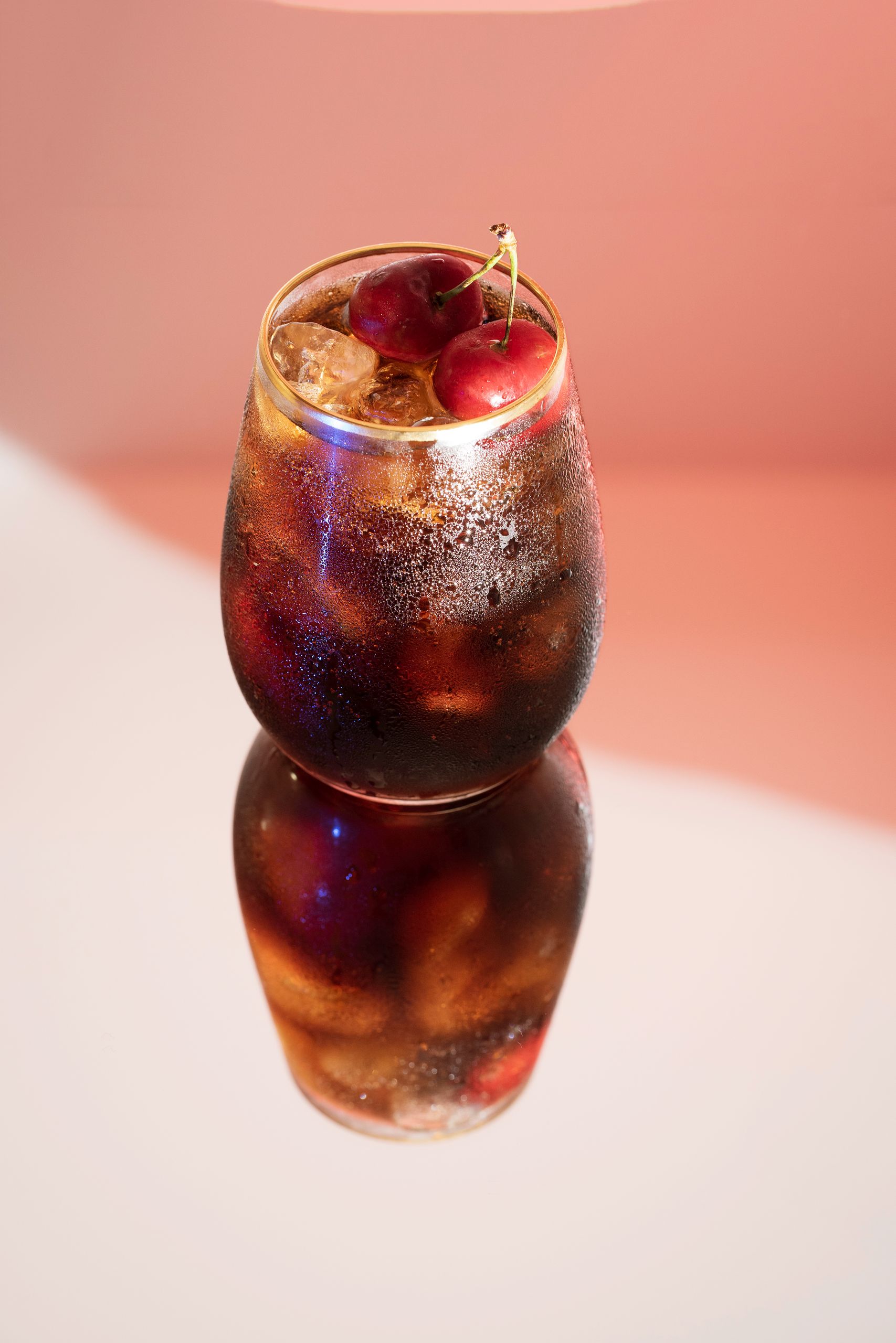 A cold glass of cherry cola with ice, featuring a vivid reflection and cherries on top.